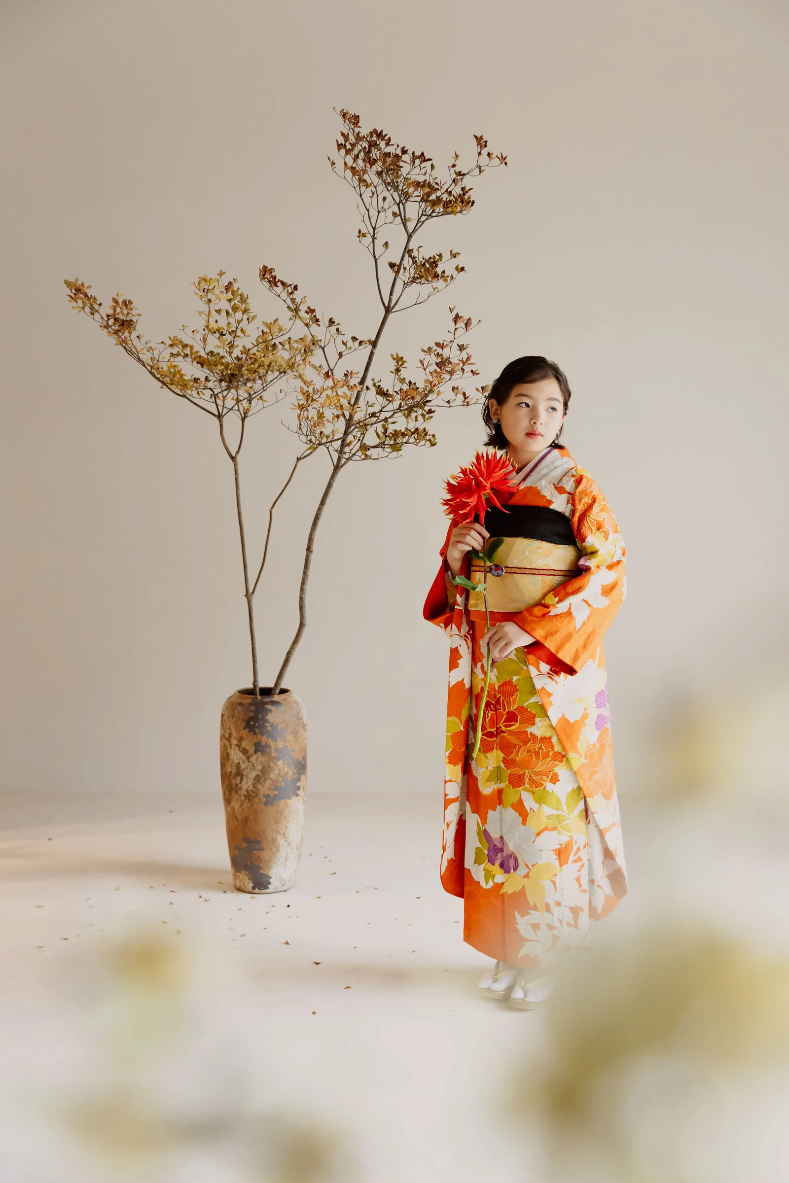 オレンジ地に橙や黄の花柄を染めた振袖を着た女児が、黒地の帯を締めて赤いダリアを手に立つ全身像。左には枝物を生けた陶製花瓶が配される。