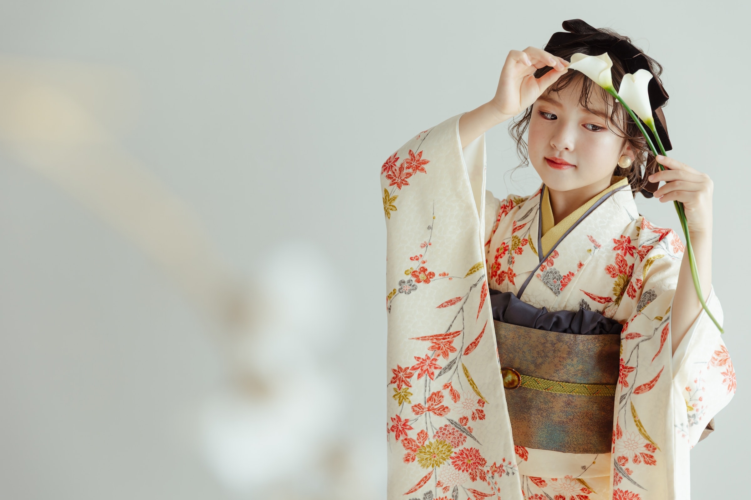 白地に赤や金の紅葉・草花を描いた振袖姿の女性が、光沢のある帯を締め、白いカラーの花を手にうつむく右寄り構図のポートレート。