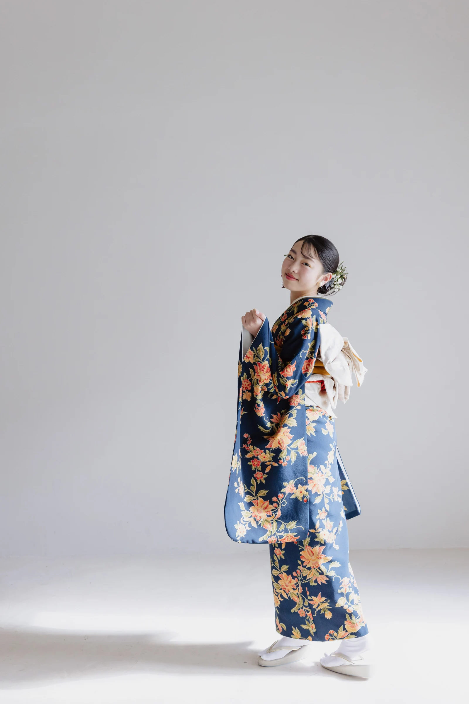 藍色地にオレンジや黄の花柄が描かれた振袖姿の女児——白い帯と髪飾りを添え、横向きに振り返り微笑む全身像、無地の明るい背景。