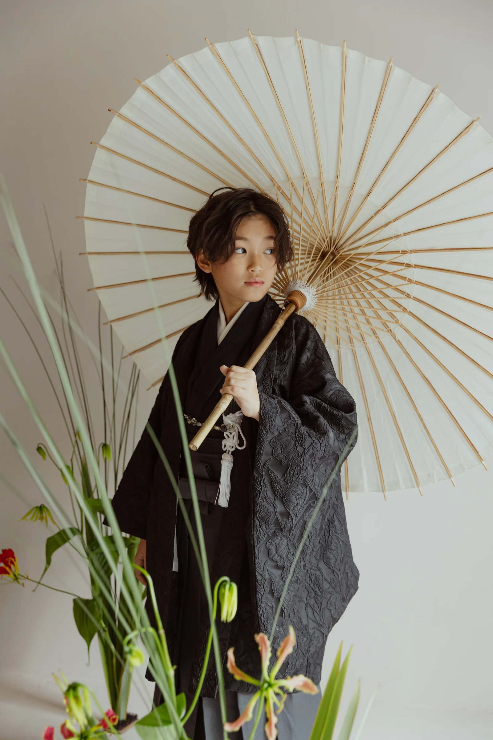 黒地に立体的な花文様の羽織と袴を着た男児が、白い和傘を肩に掛け横を向いて立つ。手前に緑の草花が配され、淡い背景の縦構図。