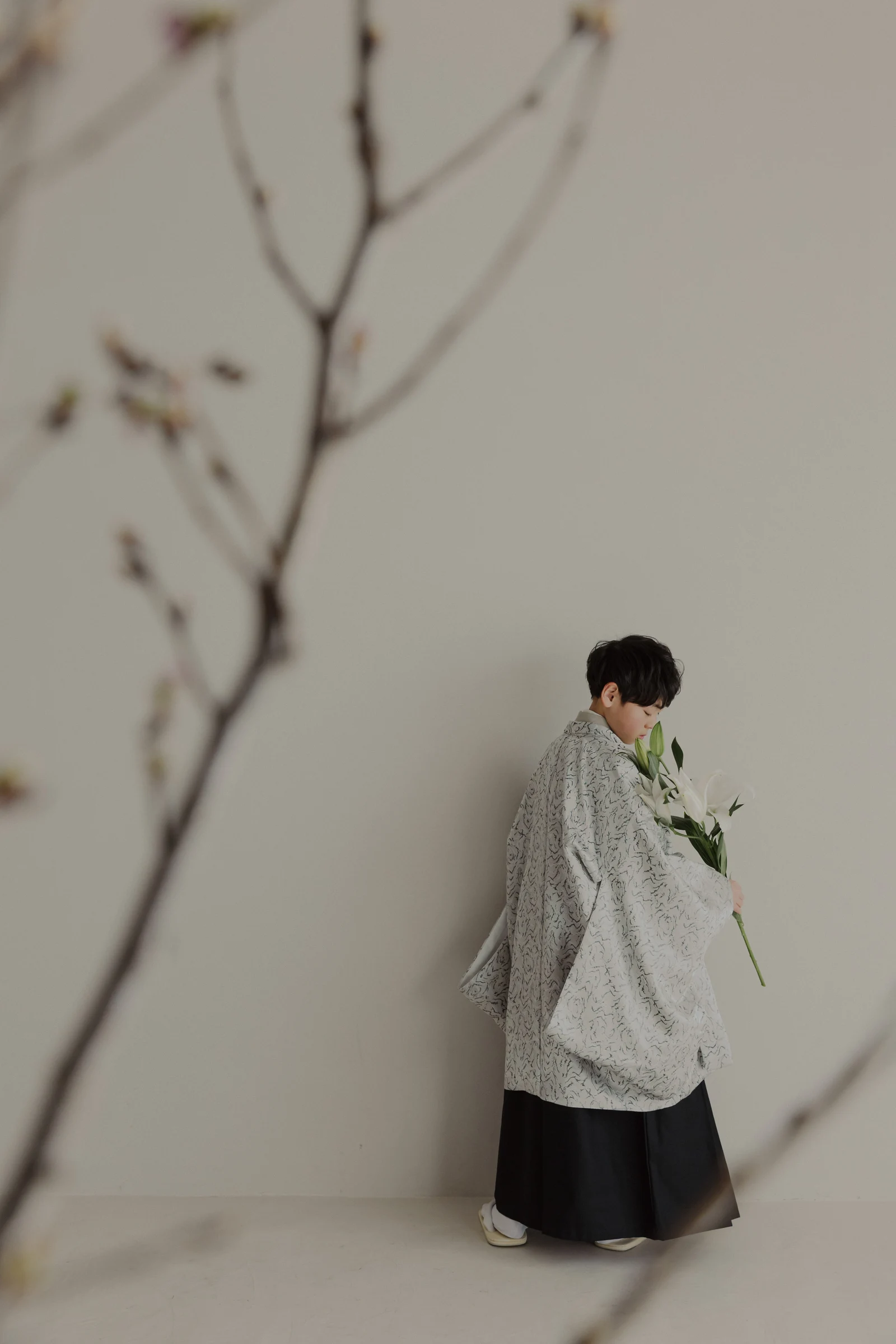白い百合の花束を持つ男児 — 細かな線模様の灰色の羽織に黒い袴を合わせ、うつむいた横向きの全身。前景に花芽の枝、淡いベージュの背景。