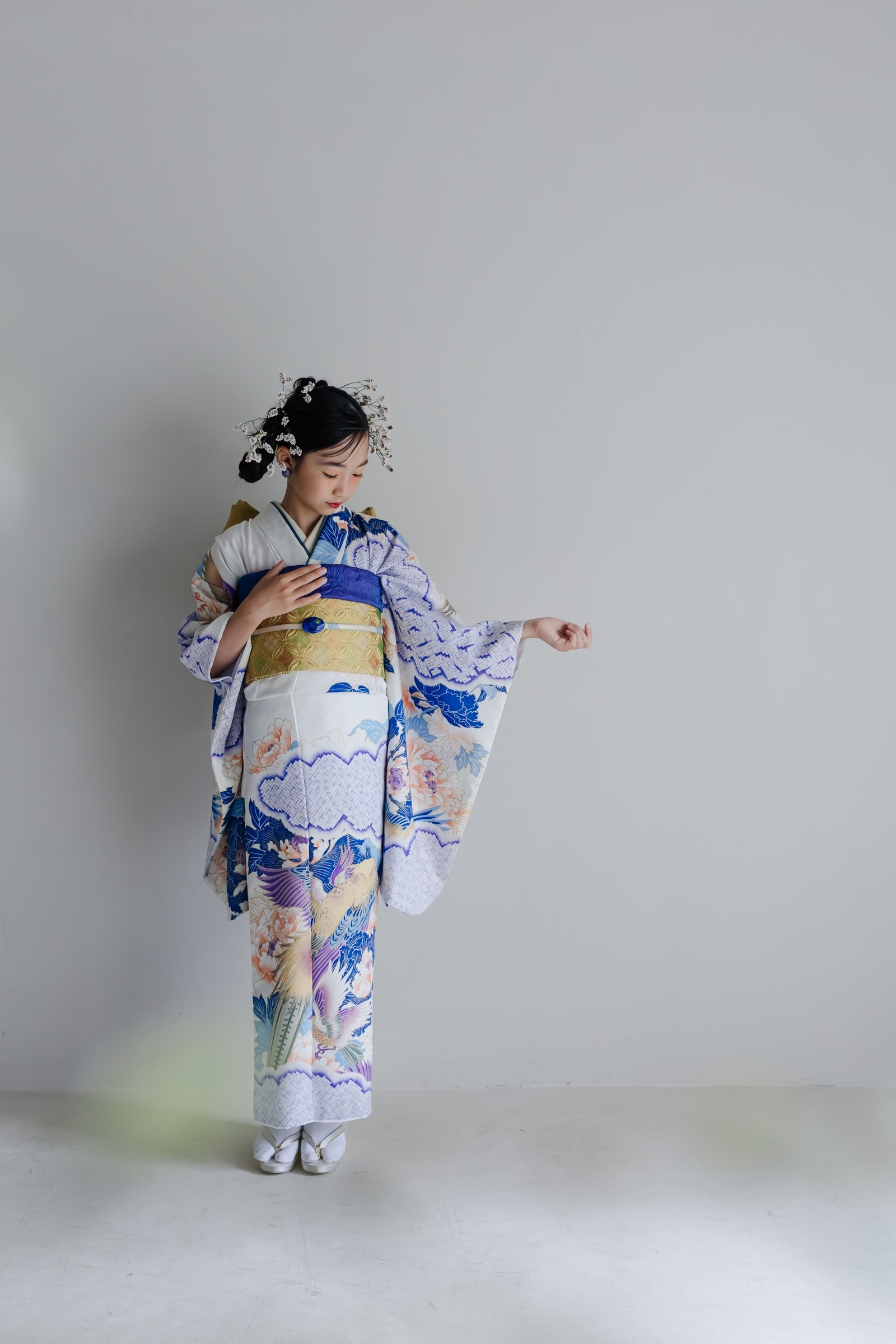 白地に青の花鳥や牡丹を配した振袖 — 金の帯と髪飾りを合わせた女性が袖を広げ、うつむいて立つ全身構図。