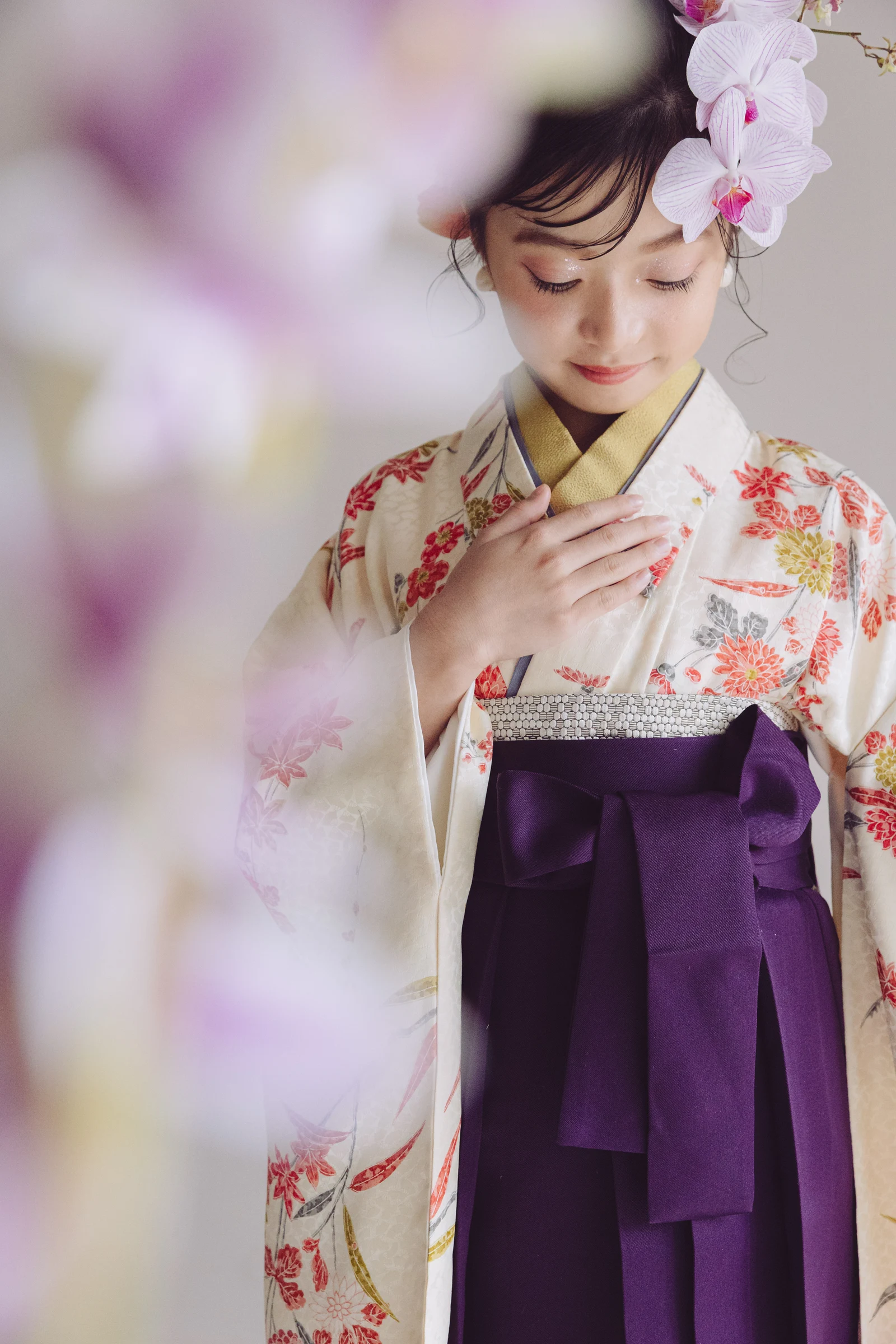 クリーム地に赤や橙の花柄が描かれた着物と紫の袴を合わせた女児、髪に淡紫の蘭を飾り胸に手を添えて伏し目で立つ姿、手前に淡いピンクの花がぼけて配される構図。