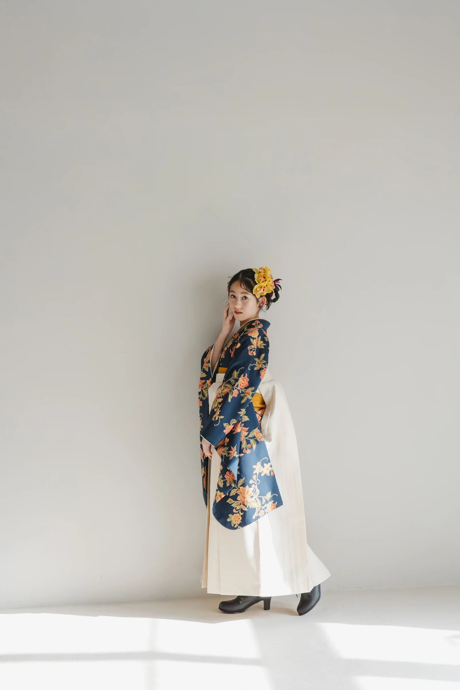 紺地に橙の花柄を配した着物に生成色の袴を合わせた女児、黄色い花飾りの結い髪。振り返る全身の横向きの立ち姿、明るい背景。