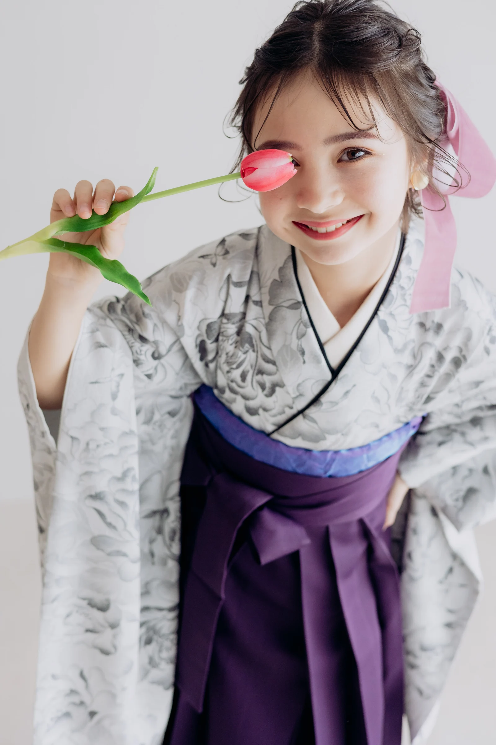 淡い灰色に墨絵調の花柄を配した着物に深紫の袴を合わせた女児が、ピンクのチューリップを片目に添え微笑む上半身の構図。髪に淡いピンクのリボン。