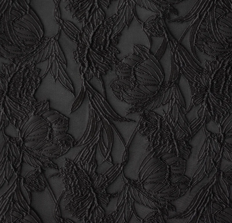 黒地に同色の花と葉の文様を立体的に織り出した生地のクローズアップ — 画面全体に密に広がる刺繍状の意匠で、陰影により凹凸が浮かび上がる構図。