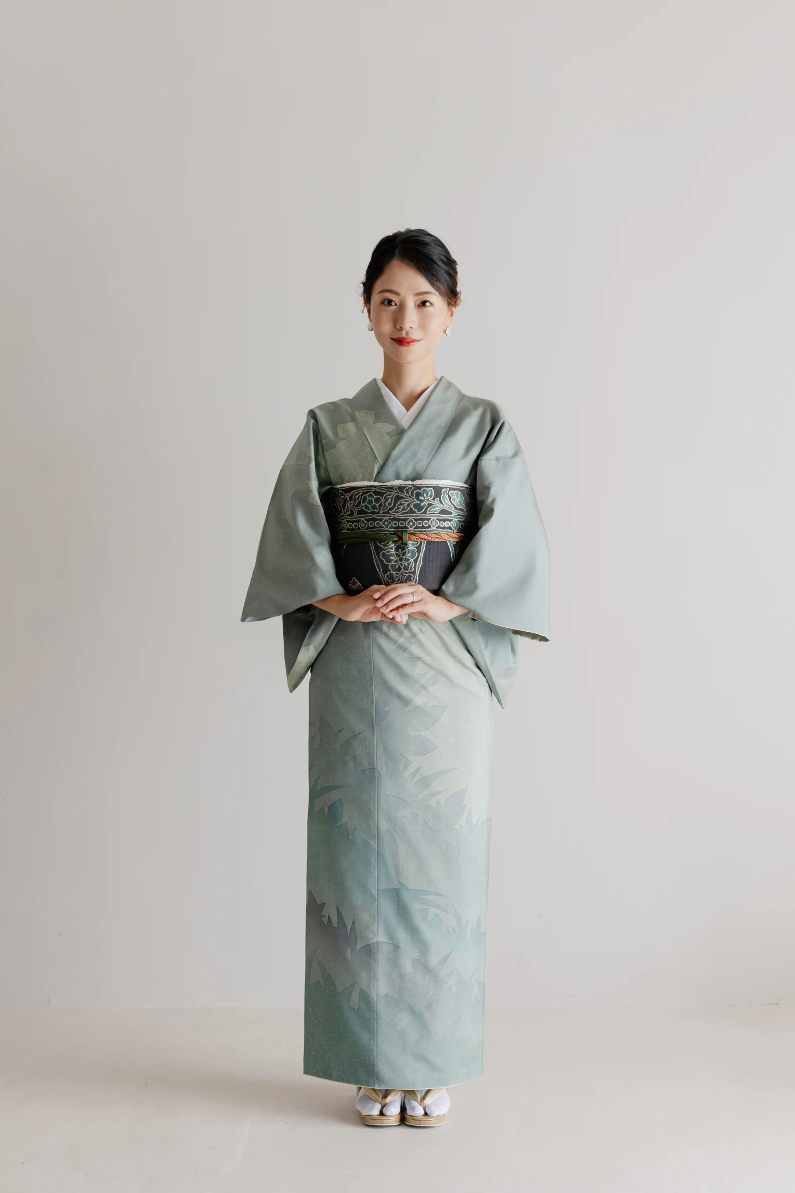 青緑系の草花文様の着物に黒地花柄の帯を締めた女性の全身正面像。手を前で重ね、白足袋に草履を履き淡い背景の前に立つ構図。