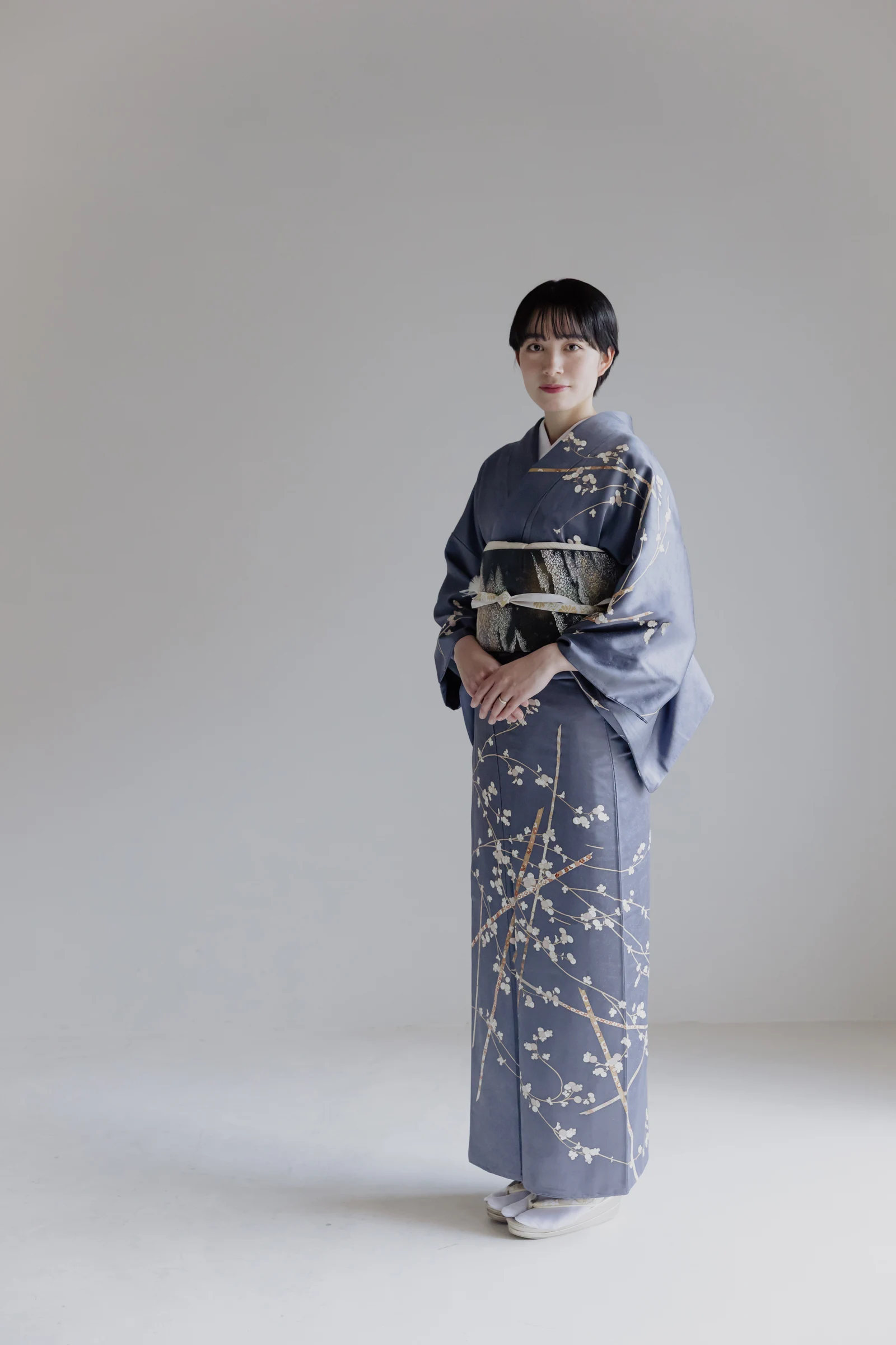 青灰色地に白い梅花と枝模様を配した着物を着た女性が、黒系の帯を締めて正面を向き両手を前で重ねて立つ全身像。背景は無地のグレー。