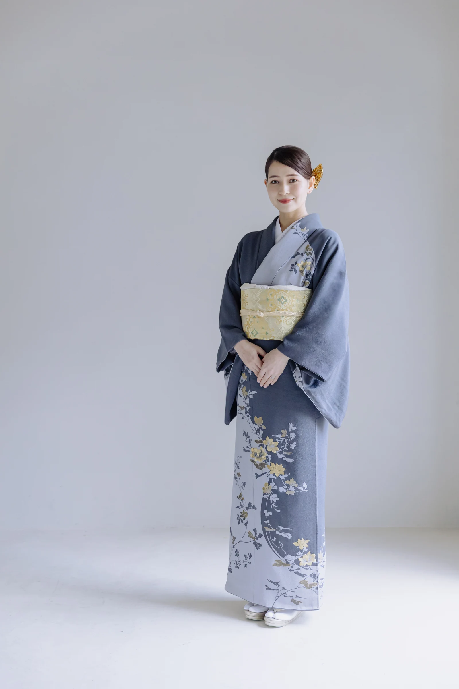 グレーから淡青へぼかした地に黄色や白の草花文様を裾と肩に配した着物姿の女性が、金色の帯を締め正面で両手を前に立つ全身像。背景は白。