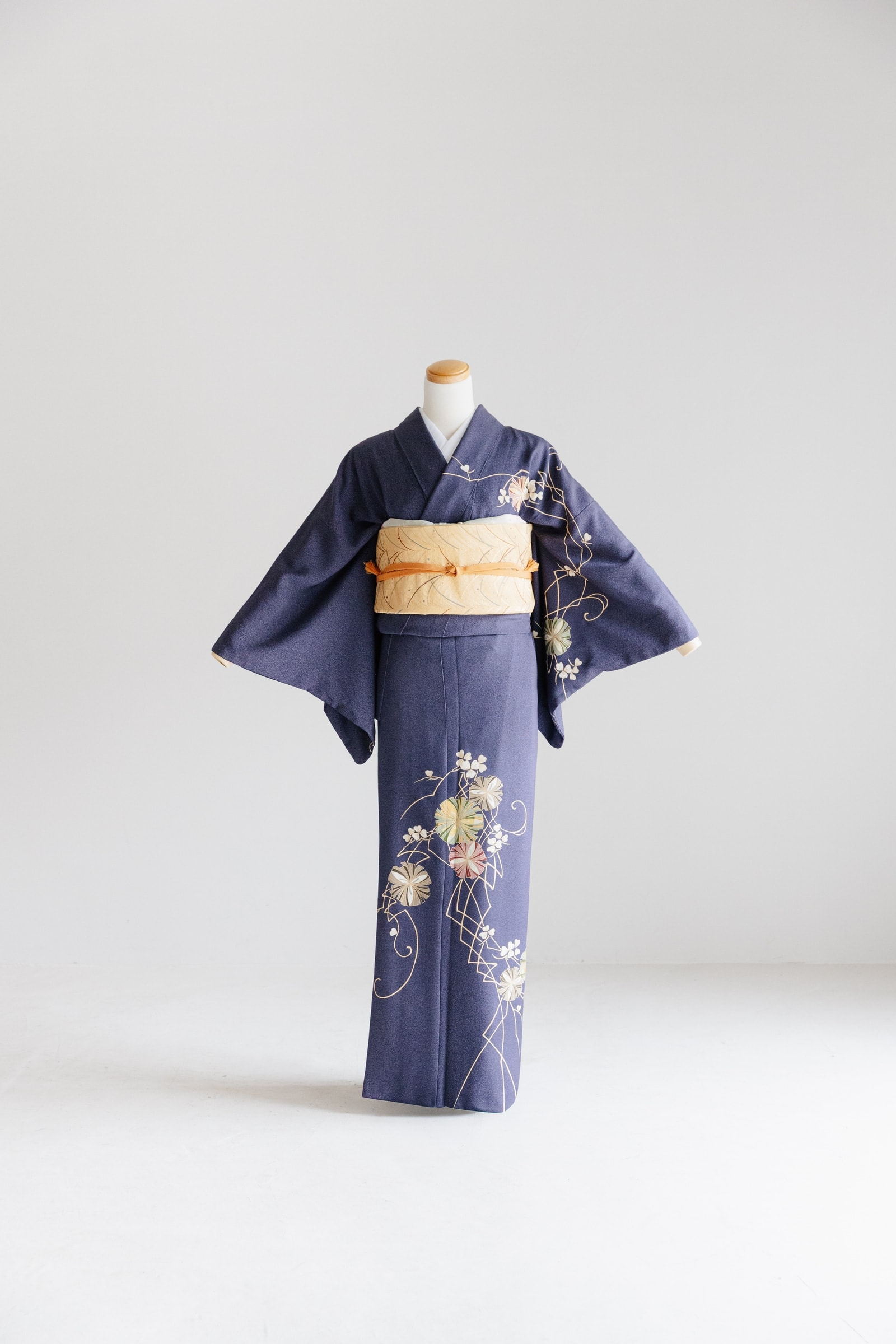 紺青の着物 — 裾と袖に菊や花の丸文を配し、淡黄色の帯と橙色の帯締めを合わせたトルソー着装の正面全身、無地背景。
