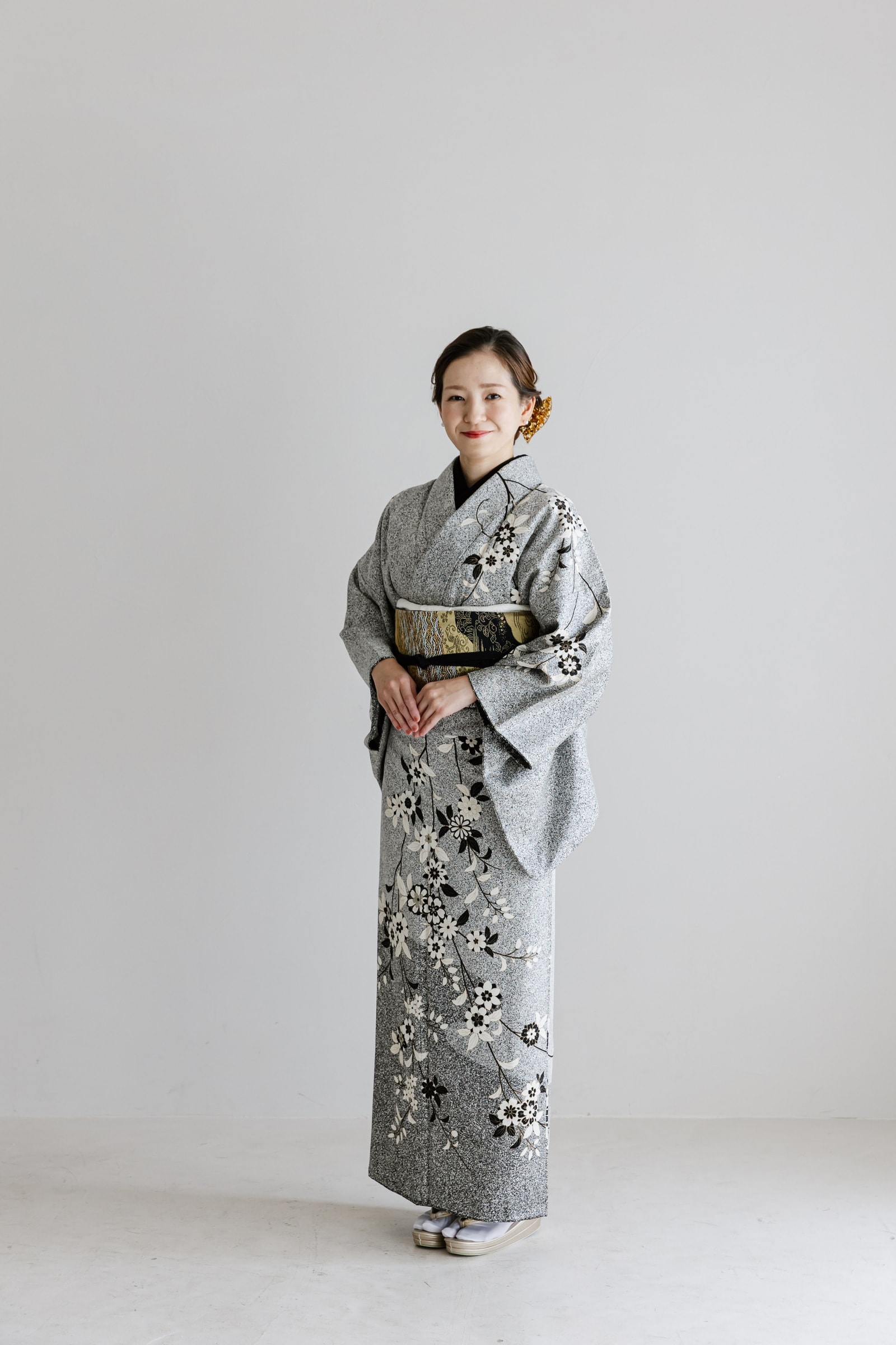 グレー地に白と黒の花柄をあしらった着物姿の女性 — 金黒の帯を締め、髪をまとめ橙色の髪飾りを付け、手を前で組んで正面に立つ全身構図。