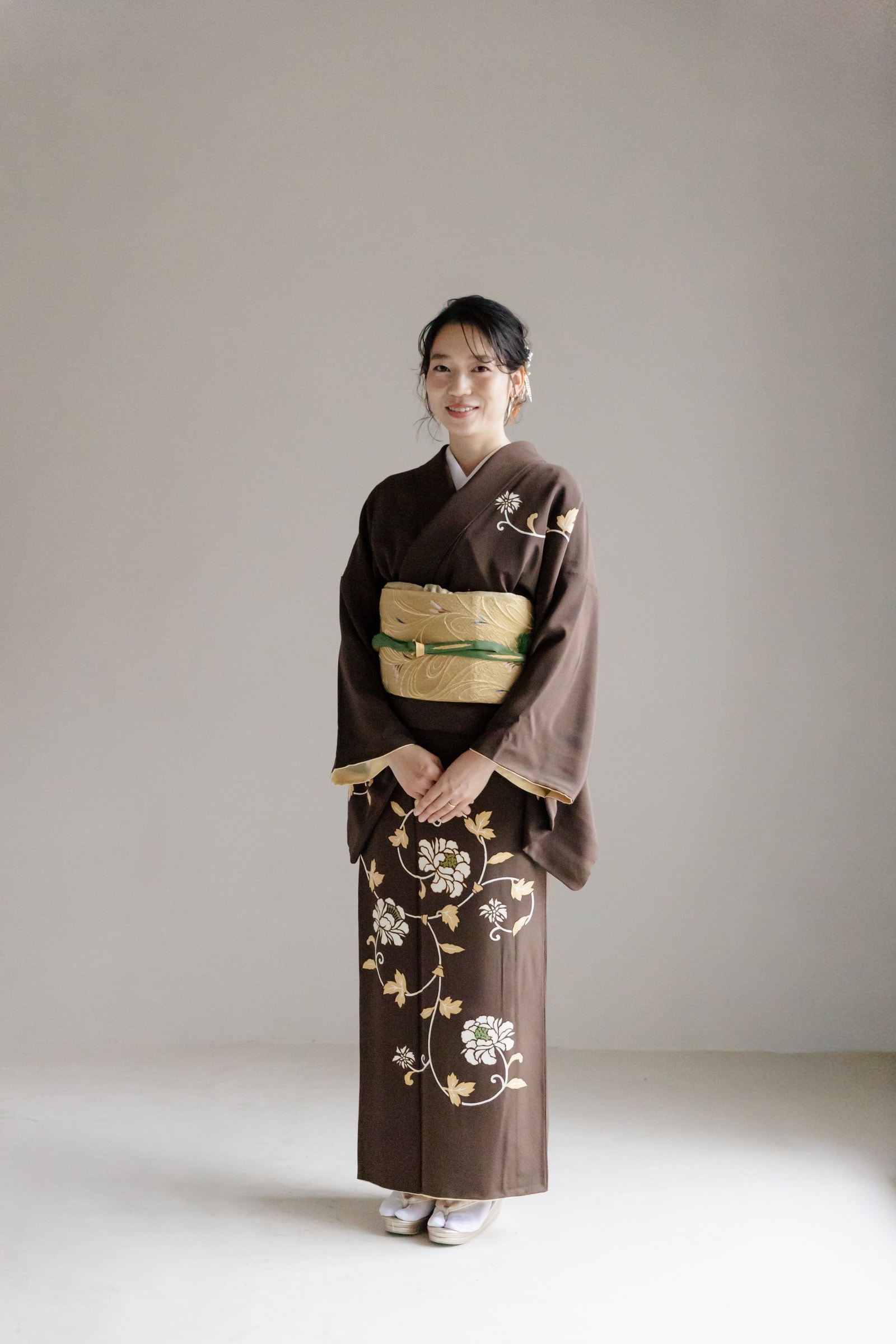 茶色地に白と金の花唐草文様を裾と肩に配した着物を着た女性の全身像。金色の帯に緑の帯締めを合わせ、白い草履を履いて立つ正面構図。