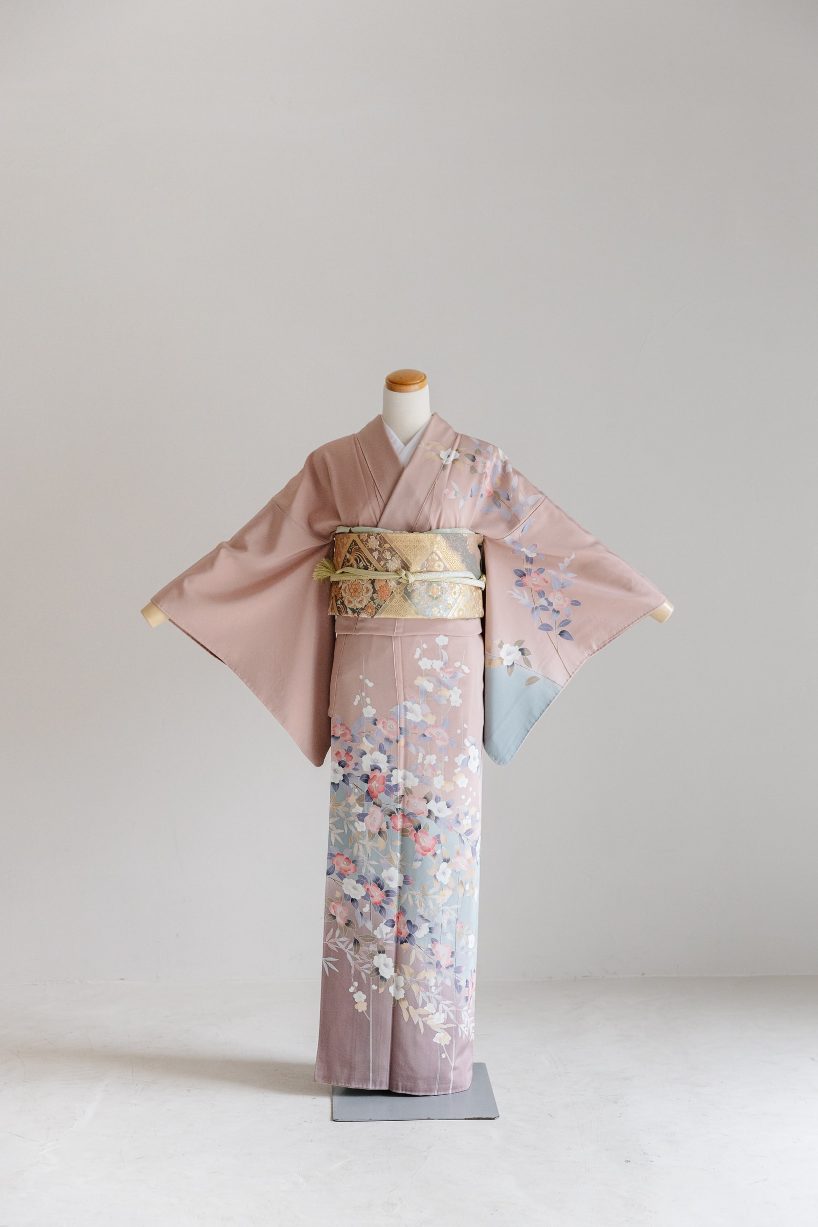 くすんだピンク地に裾へ向かって水色のぼかしと白・青・桃色の花柄を配した訪問着 — 金襴の袋帯を締めトルソーに正面から着付けた全身像。