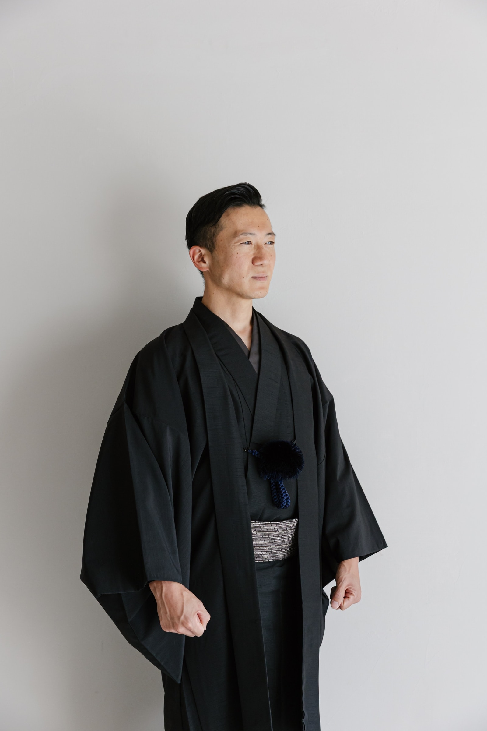 黒の着物と羽織を着た男性が横を向いて立つ姿。羽織紐は紺色の房付き、帯は白黒の細かな柄。灰色の無地背景で上半身を写した構図。